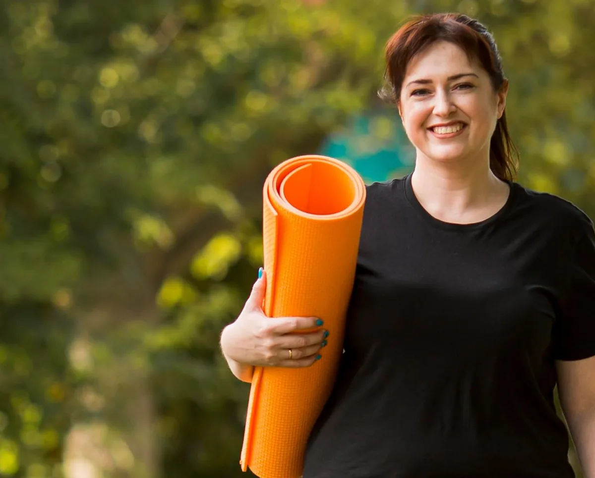 Smiling woman carrying an orange yoga mat outdoors
